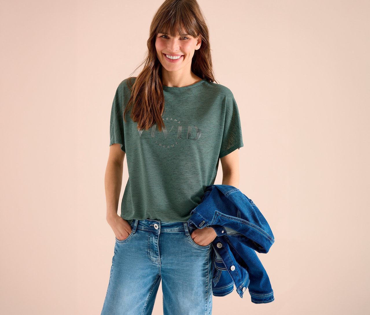 Femme souriante aux cheveux bruns portant un t-shirt vert imprimé, un jean bleu et une veste en jean sur le bras sur fond beige.