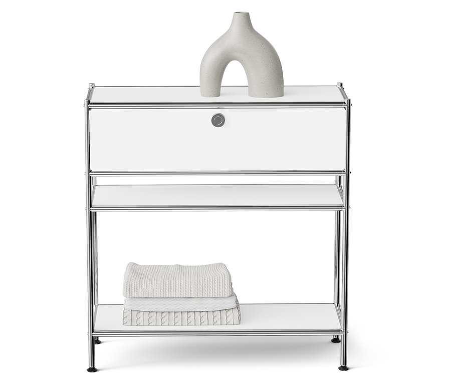 Une table de chevet blanche avec un cadre chromé avec un vase sculptural gris sur le dessus et des serviettes pliées sur une étagère.