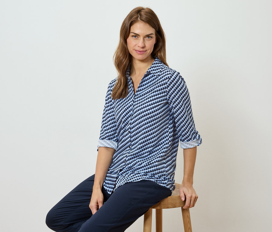 Une femme est assise sur un tabouret, portant un chemisier à motifs bleu et blanc et un pantalon foncé.