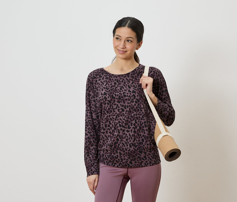 Femme portant un haut léopard violet et noir et un legging violet, tenant un tapis de yoga roulé sur l'épaule.