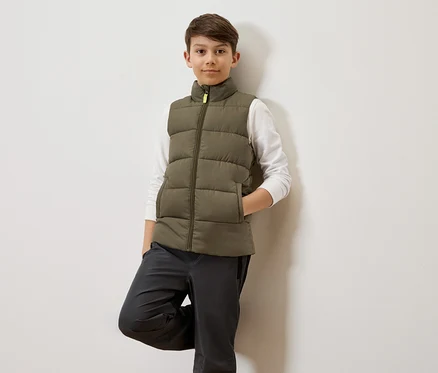 Un garçon aux cheveux bruns s'appuie contre un mur blanc, portant un gilet matelassé vert olive sur une chemise blanche à manches longues et un pantalon gris.