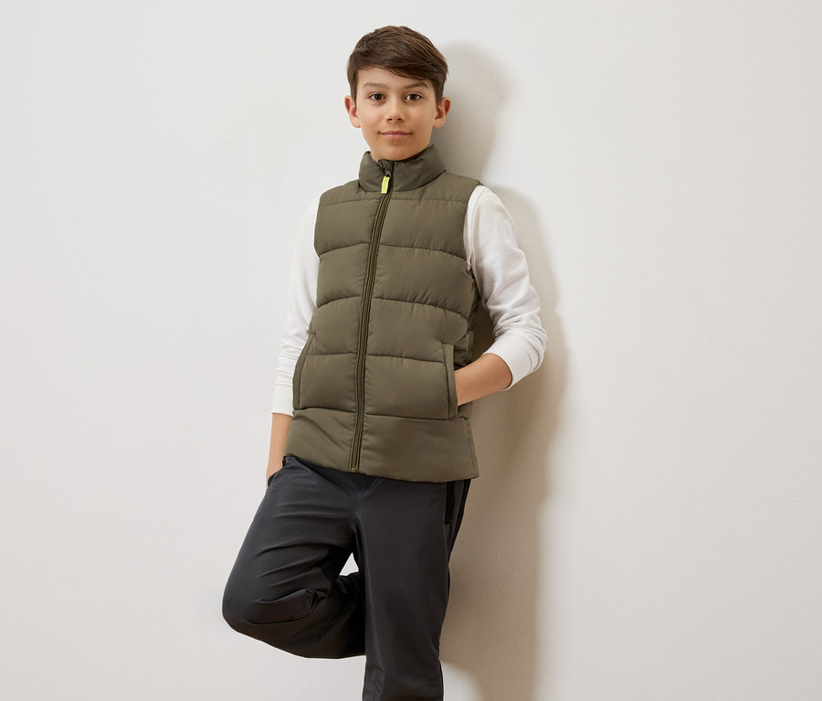 Un garçon aux cheveux bruns s'appuie contre un mur blanc, portant un gilet matelassé vert olive sur une chemise blanche à manches longues et un pantalon gris.