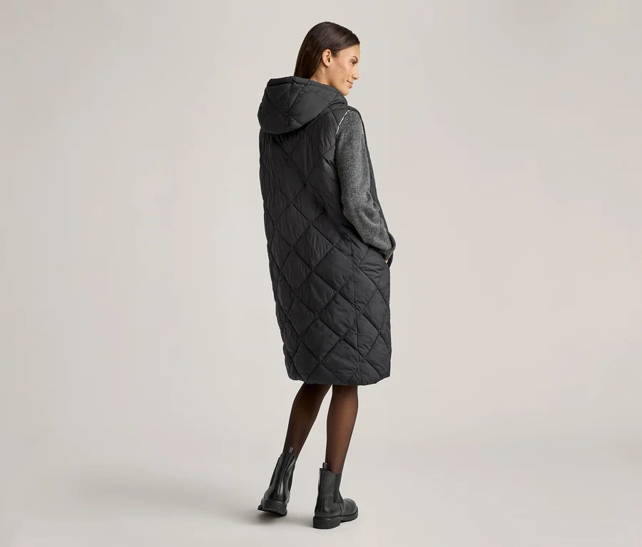 Femme vue de dos portant une longue veste sans manches noire à capuche, un pull gris, des collants et des bottines.
