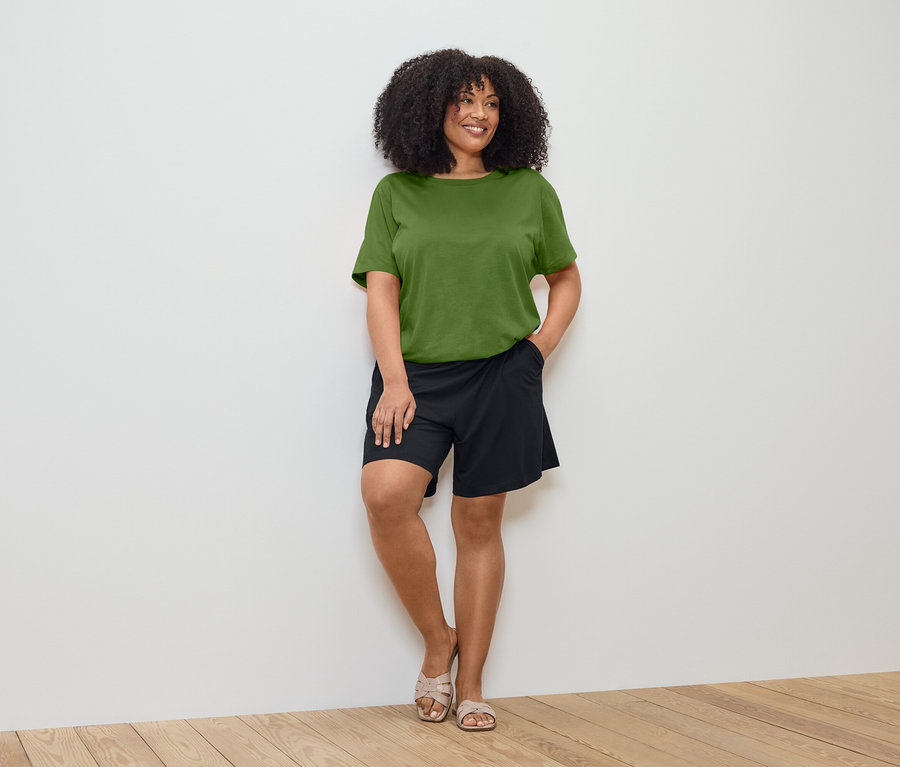 Une femme aux cheveux bouclés, portant un t-shirt vert olive et un short noir, est appuyée contre un mur blanc.