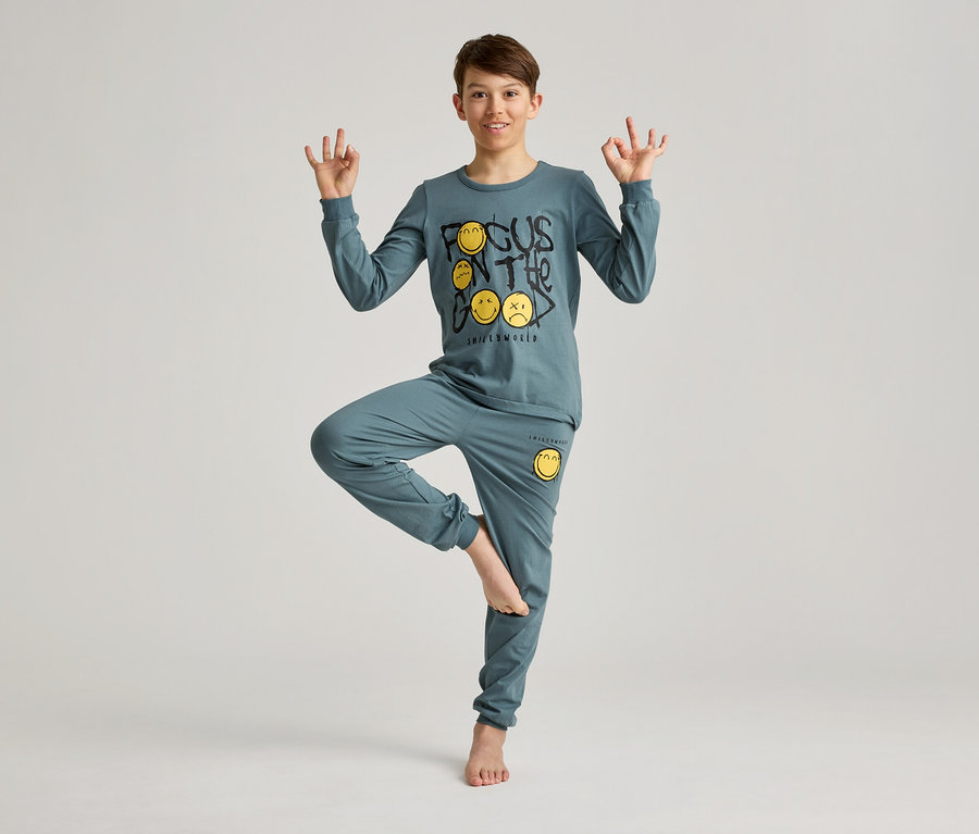 Un garçon en pyjama bleu avec un motif de smiley, en équilibre sur une jambe et faisant des mudras avec ses mains.