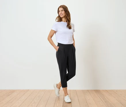 Une femme portant un t-shirt blanc et un pantalon noir pose devant un mur blanc.