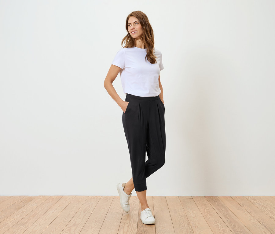 Une femme portant un t-shirt blanc et un pantalon noir pose devant un mur blanc.