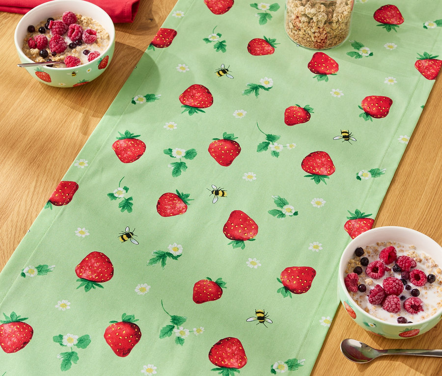 Sur une table en bois se trouve un chemin de table à motifs de fraises, dessus des bols de céréales, un verre de muesli et une bouteille de lait.