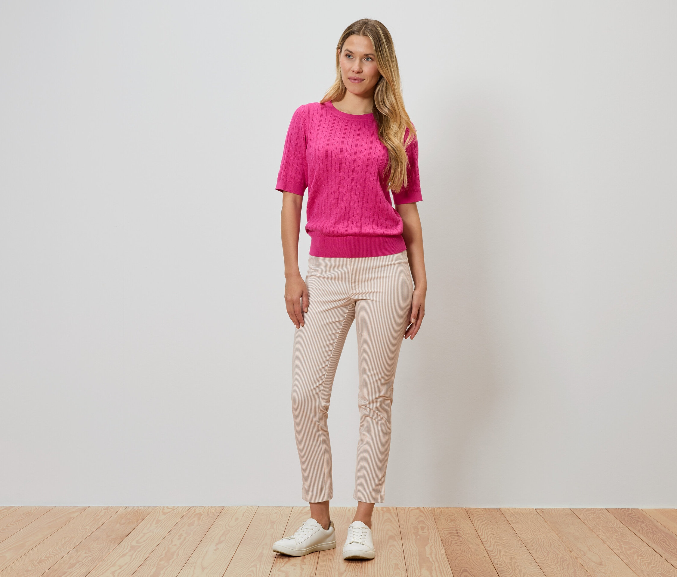 Une femme se tient devant un mur, portant un pull rose, un pantalon rayé et des chaussures blanches.