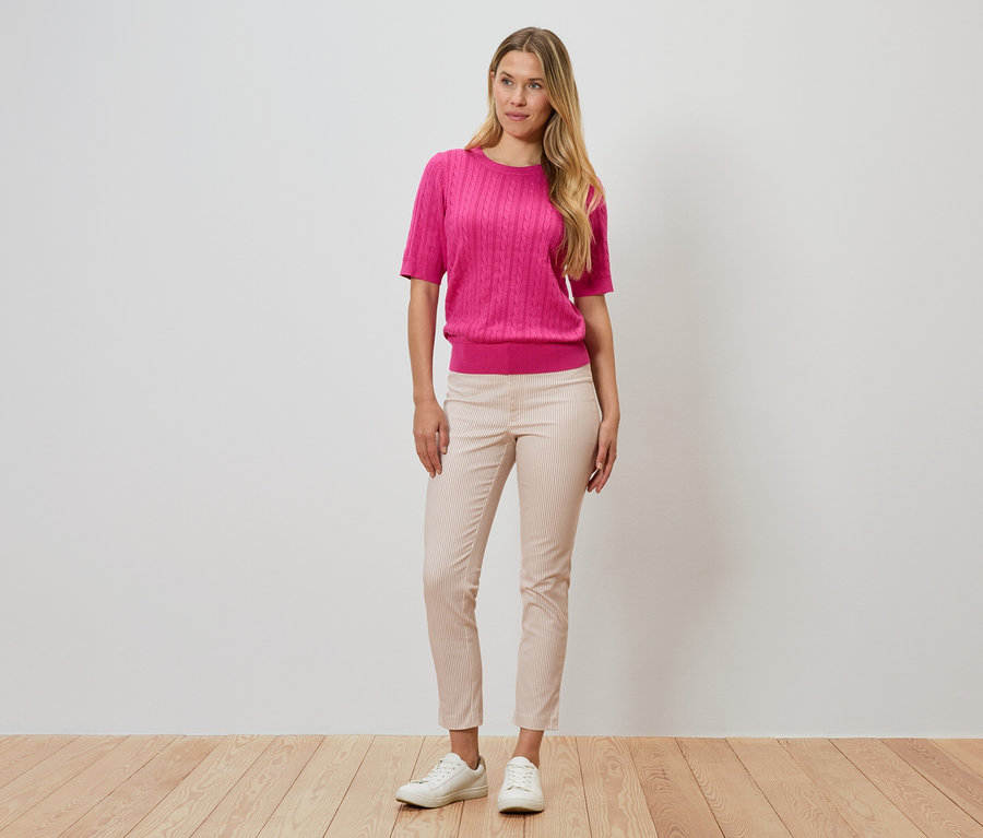 Une femme se tient devant un mur, portant un pull rose, un pantalon rayé et des chaussures blanches.