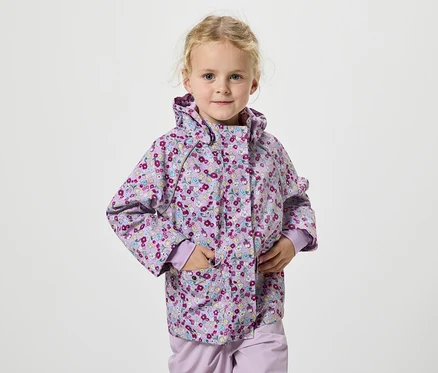 Une fillette blonde portant une veste à fleurs lilas se tient devant un fond blanc.