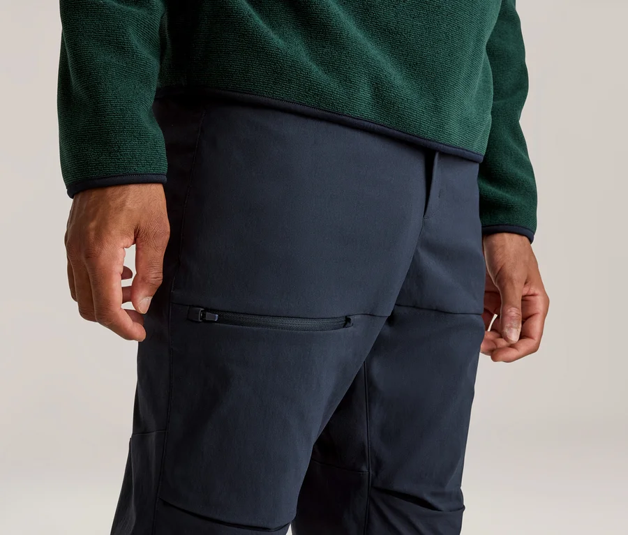 Gros plan d'une personne portant un haut polaire vert foncé et un pantalon bleu foncé avec une poche zippée sur la cuisse. Les mains sont visibles sur les côtés.