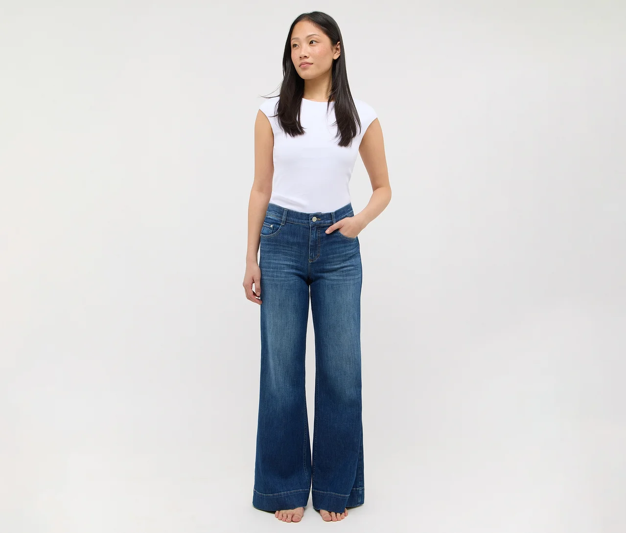 Une femme debout, vêtue d'un t-shirt blanc et d'un jean bleu à jambes larges, les mains dans les poches.
