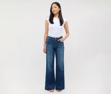 Une femme debout, vêtue d'un t-shirt blanc et d'un jean bleu à jambes larges, les mains dans les poches.