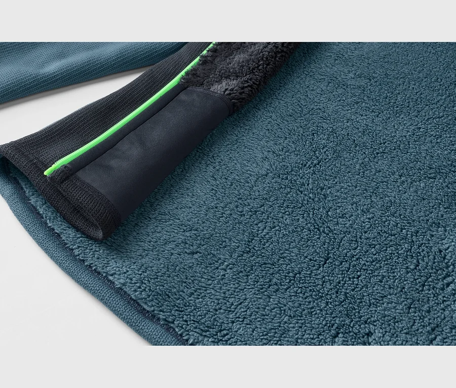Gros plan d'un tissu polaire sarcelle, montrant sa texture moelleuse. Une bordure gris foncé avec un détail de fermeture éclair vert néon traverse le tissu en diagonale.