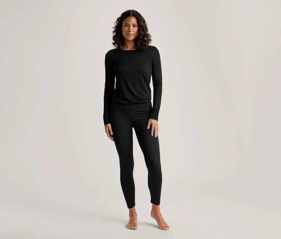Une femme aux cheveux foncés et bouclés se tient de face, vêtue d'un haut noir à manches longues et d'un legging noir. Elle est pieds nus.