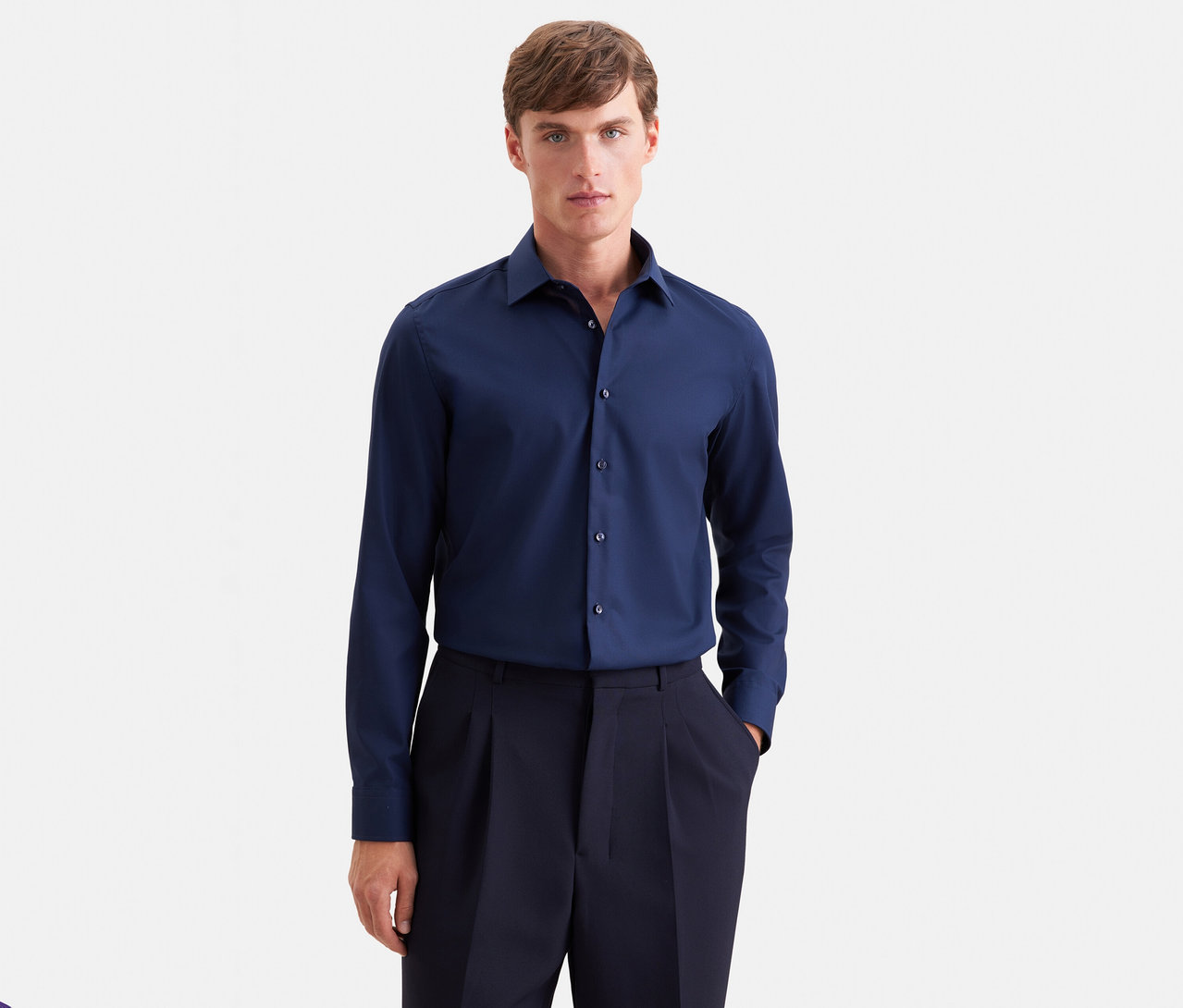 Homme portant une chemise à manches longues bleu foncé et un pantalon plissé bleu foncé, debout de face avec une main dans la poche.