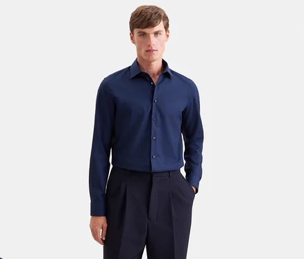 Homme portant une chemise à manches longues bleu foncé et un pantalon plissé bleu foncé, debout de face avec une main dans la poche.