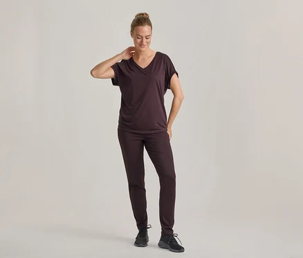 Femme portant un t-shirt brun foncé à col en V et un pantalon assorti, avec des baskets noires, debout, regardant vers le bas, une main derrière la nuque.