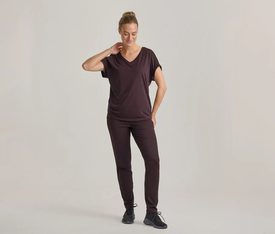 Femme portant un t-shirt brun foncé à col en V et un pantalon assorti, avec des baskets noires, debout, regardant vers le bas, une main derrière la nuque.