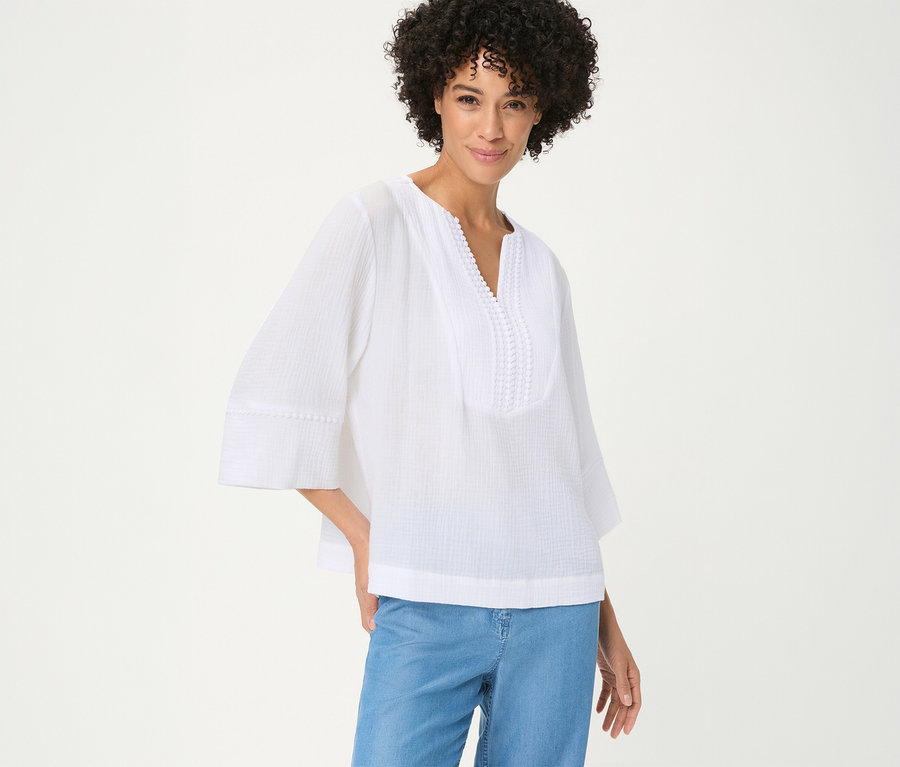 Femme souriante aux cheveux bouclés portant une blouse tissée Olsen blanche à col en V avec broderie et manches 3/4, et un jean bleu.