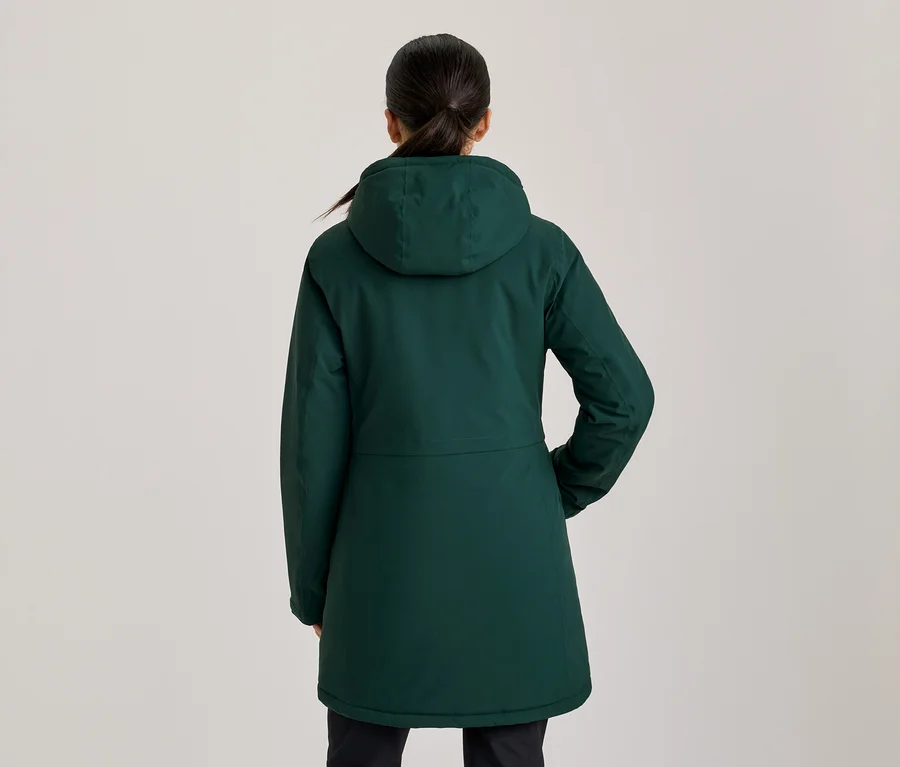 Femme vue de dos, portant un manteau à capuche vert foncé, la main droite dans une poche. Ses cheveux foncés sont attachés en queue de cheval.