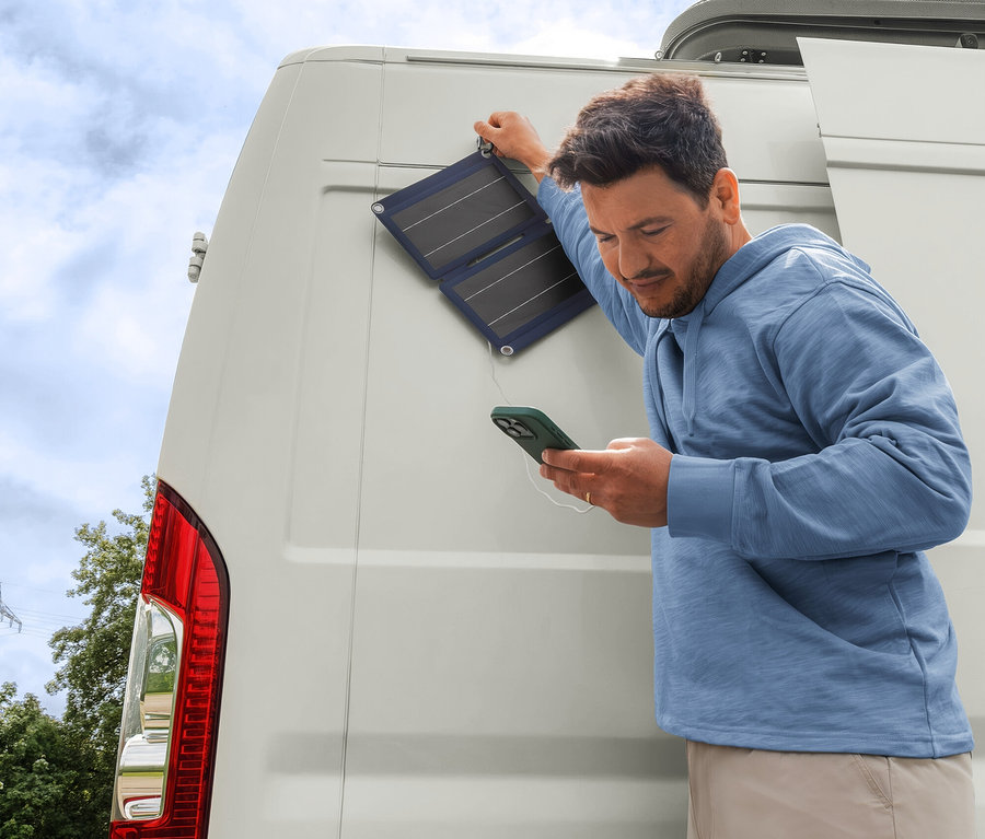 Homme en sweat-shirt à capuche et short beige chargeant son téléphone avec un chargeur solaire pliable de 10 W fixé à un camping-car blanc.