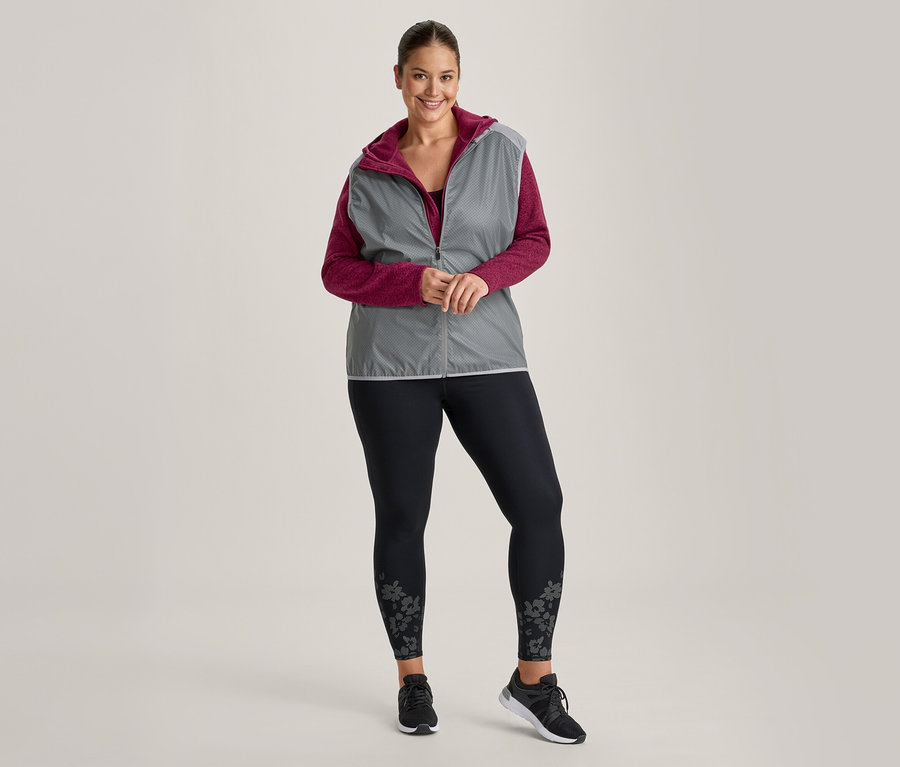 Femme souriante portant un gilet gris à capuche sur un haut magenta à manches longues, un legging noir à motifs floraux et des baskets noires.
