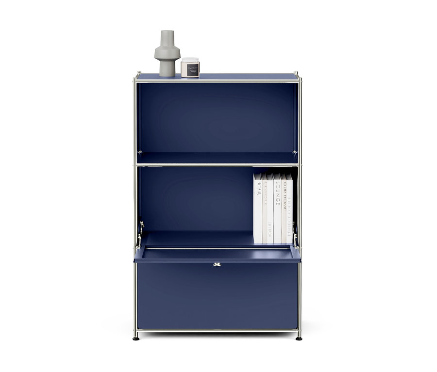 Armoire modulaire bleu foncé avec cadre chromé et trois sections. Sur le dessus, un vase gris et une bougie blanche. La section centrale contient une pile de livres blancs. La section inférieure a une porte abattante partiellement ouverte au-dessus d'un tiroir fermé.