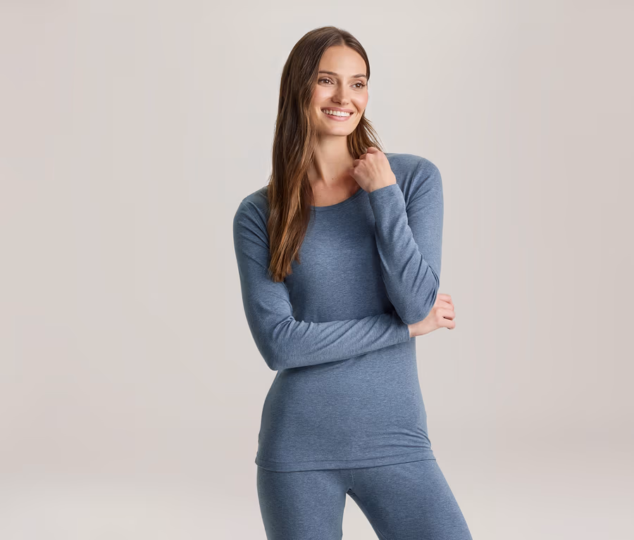 Une femme souriante aux longs cheveux bruns porte un haut bleu à manches longues et un legging assorti, regardant vers la droite.