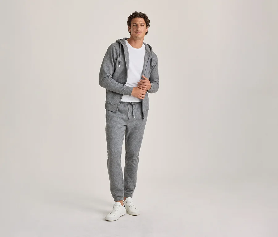 Un homme portant un sweat à capuche gris, un t-shirt blanc et un pantalon de jogging gris, avec des baskets blanches, regarde la caméra.