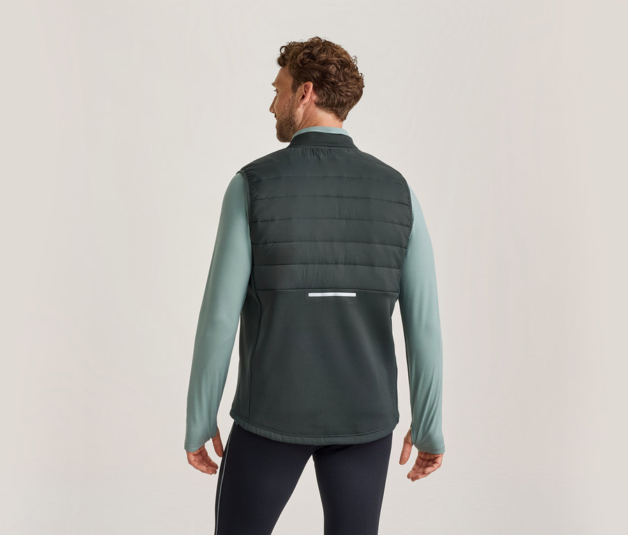 Homme vu de dos, portant un gilet matelassé vert foncé sur un t-shirt à manches longues vert clair et un pantalon foncé. Le gilet a une bande réfléchissante au dos.