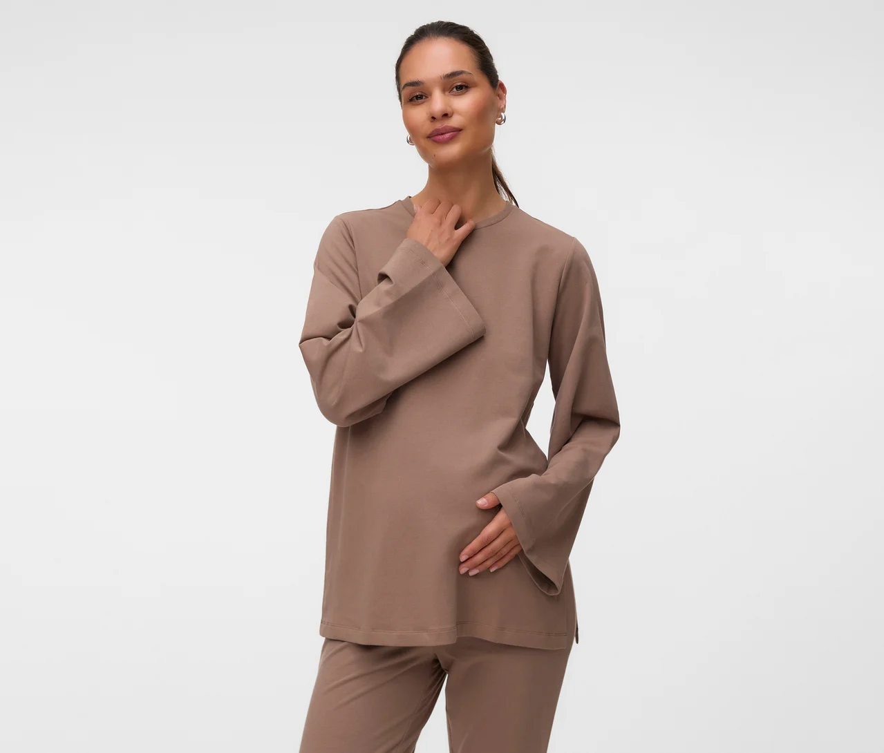 Une femme enceinte porte un sweat-shirt MAMALICIOUS « MLSINE LS » marron clair et un pantalon assorti, sa main droite sur son cou et sa main gauche sur son ventre.