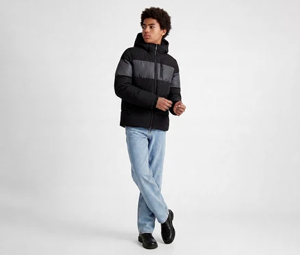 Jeune homme aux cheveux bouclés portant une doudoune rayée noire et grise, un jean bleu clair et des chaussures noires, debout les jambes croisées et les mains jointes.