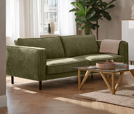 Un canapé 3 places en velours côtelé vert dans un salon, à côté d'une lampe de table en métal noire et d'un meuble haut « Bekeson » blanc.