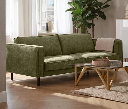 Un canapé 3 places en velours côtelé vert dans un salon, à côté d'une lampe de table en métal noire et d'un meuble haut « Bekeson » blanc.