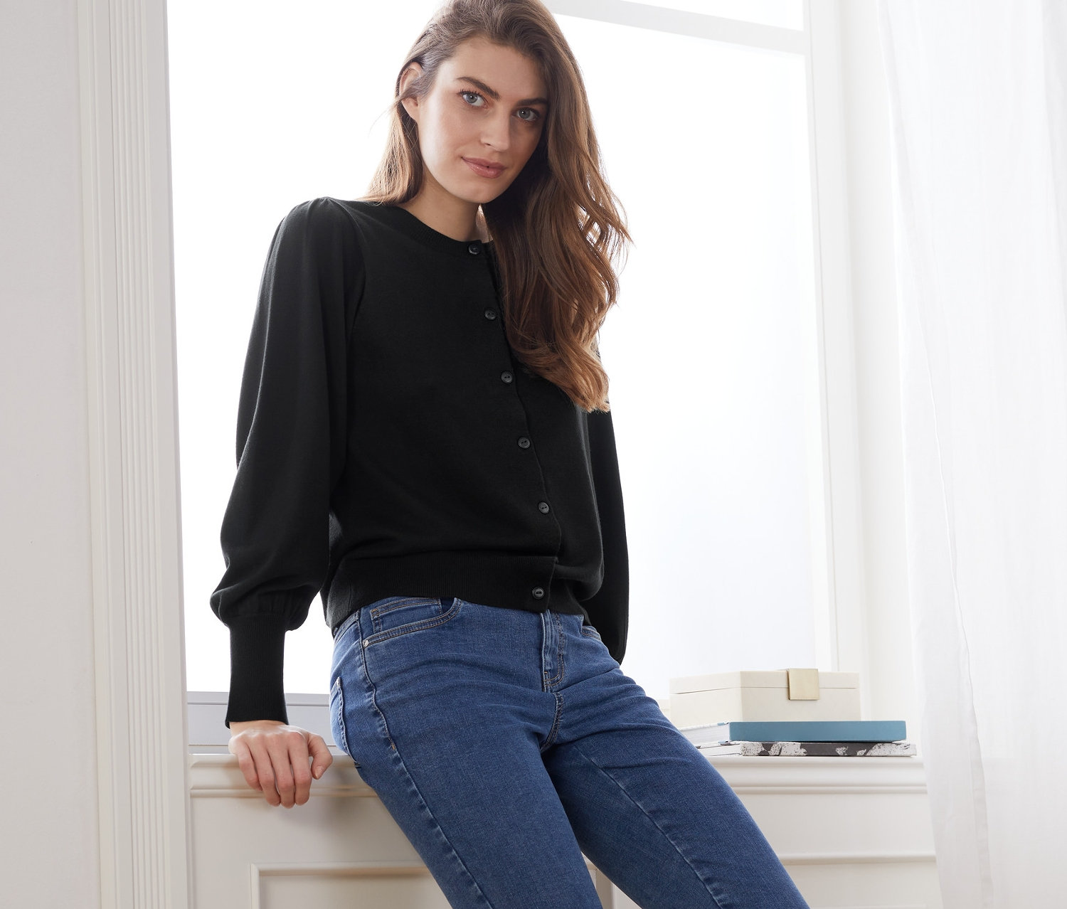 Femme assise sur un rebord de fenêtre portant une jaquette en maille fine avec des manches bouffantes et un jean bleu.