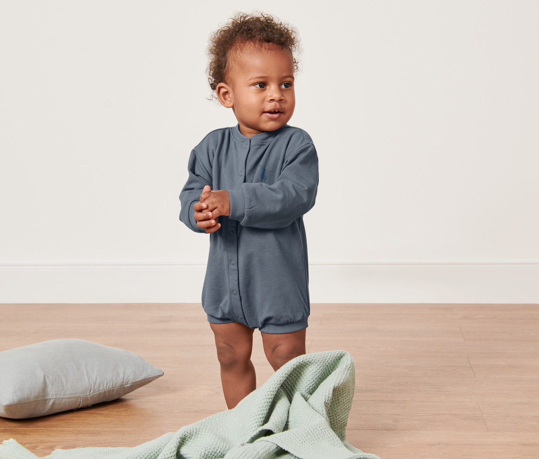 Un bébé debout sur le sol porte une barboteuse en sweat bleue.