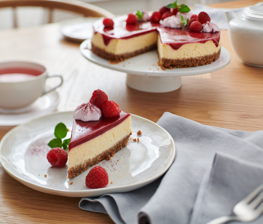 Sur une table, une assiette avec une part de gâteau décorée de framboises et de menthe. À l'arrière-plan, le reste du gâteau sur un plateau, une tasse de thé et une théière. Pelle à tarte