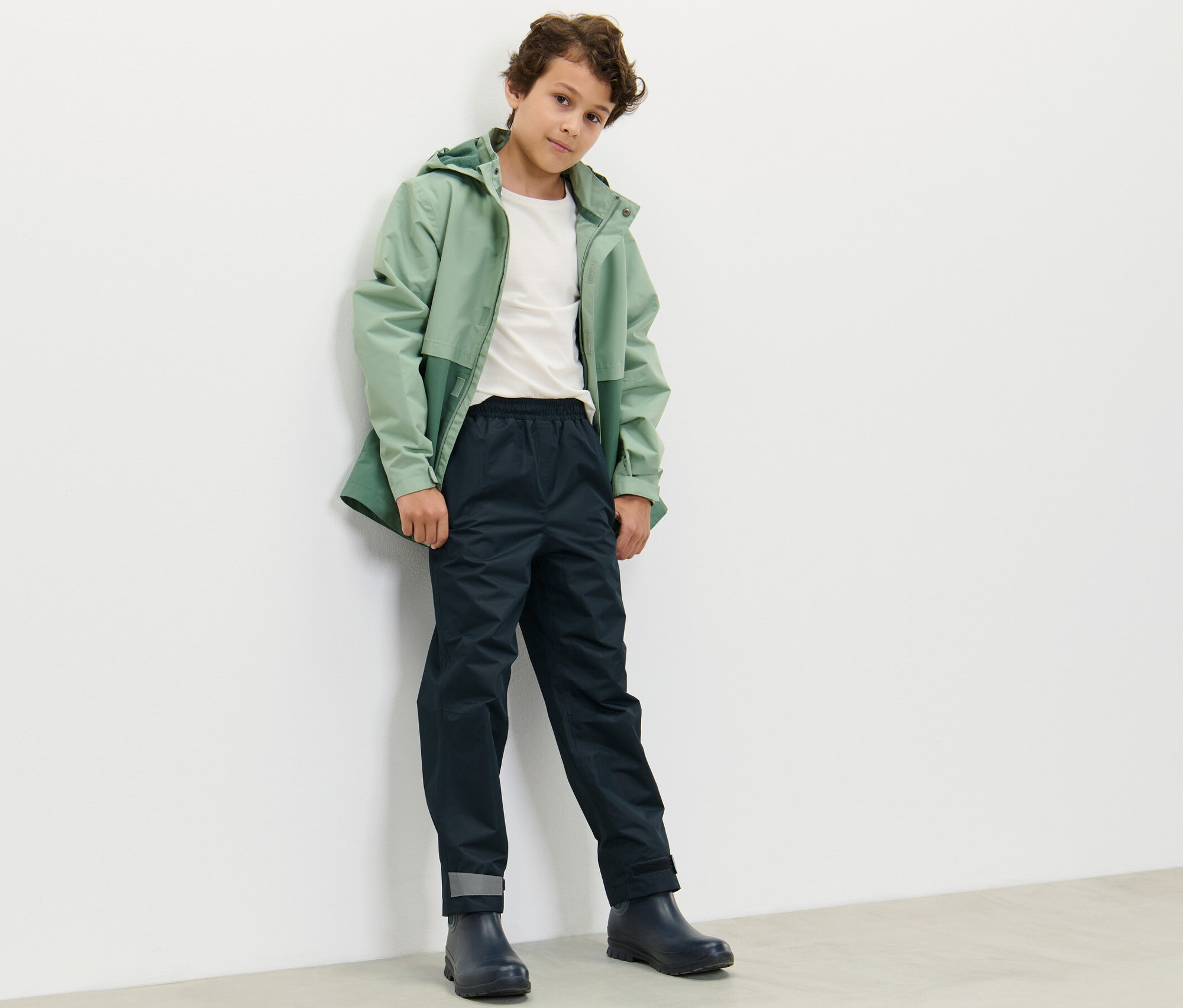 Un garçon vêtu d'une veste verte et d'un pantalon de pluie bleu marine pour enfants se tient contre un mur blanc.