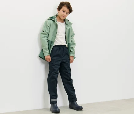 Un garçon vêtu d'une veste verte et d'un pantalon de pluie bleu marine pour enfants se tient contre un mur blanc.