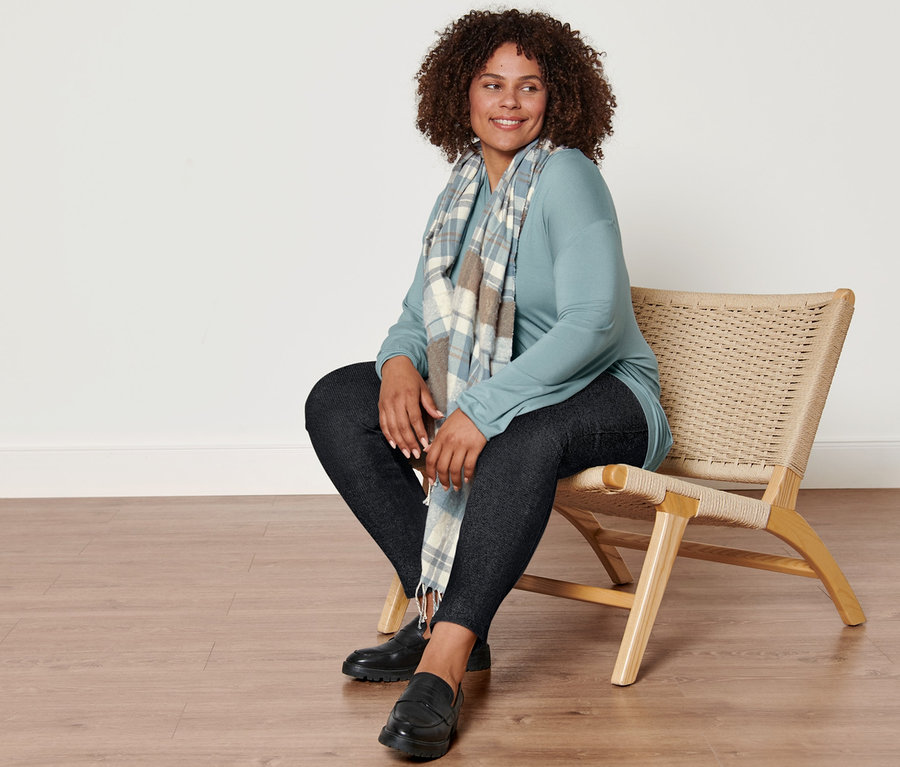 Une femme est assise sur une chaise et porte une écharpe tissée à carreaux, un legging doublé à l’aspect jean, un t-shirt manches longues avec encolure en V, bleu fumée et des loafers en cuir.