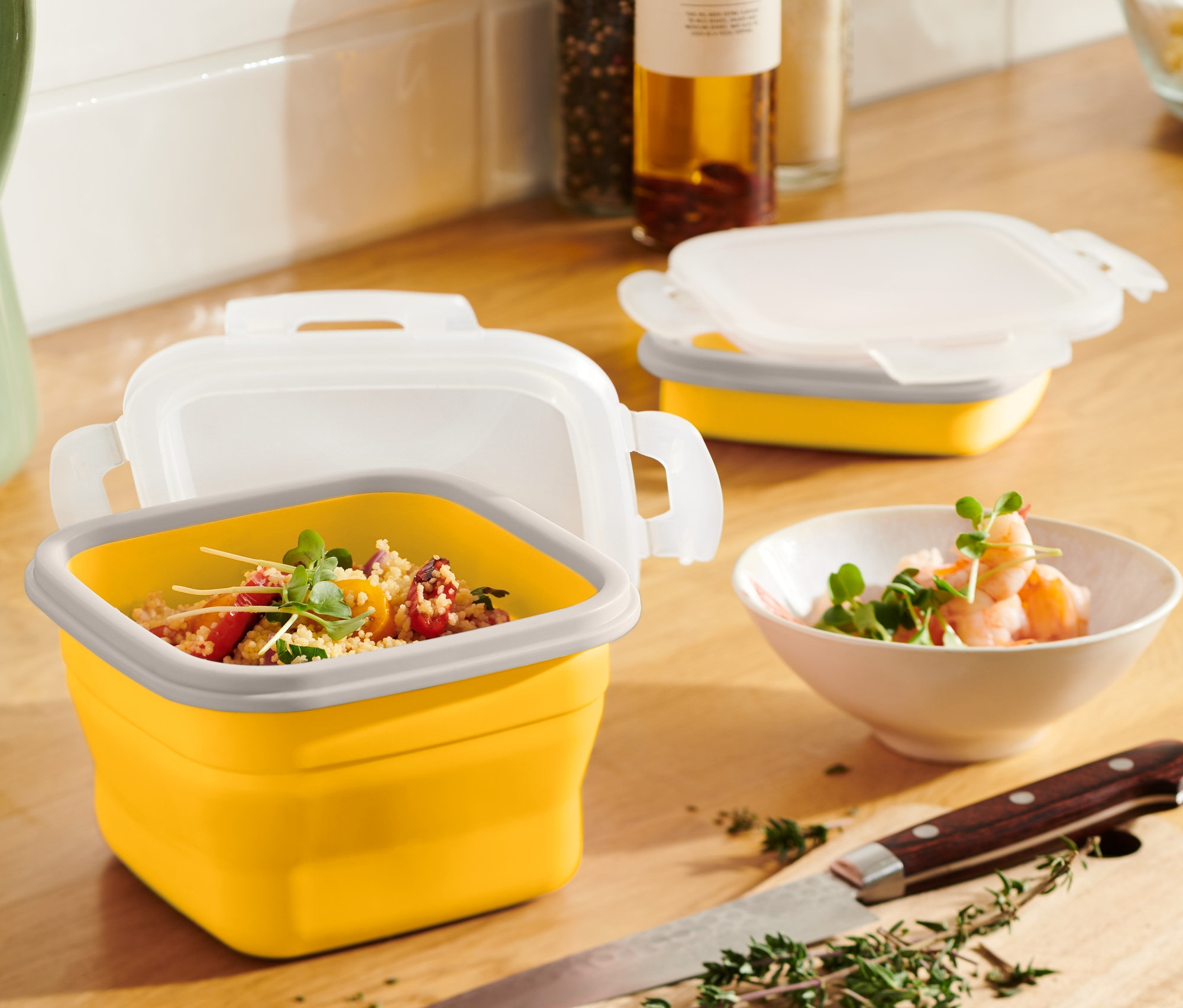 Une boîte fraîcheur pliable jaune avec du contenu est posée sur une table en bois à côté d'un pichet vert et d'un bol de crevettes.