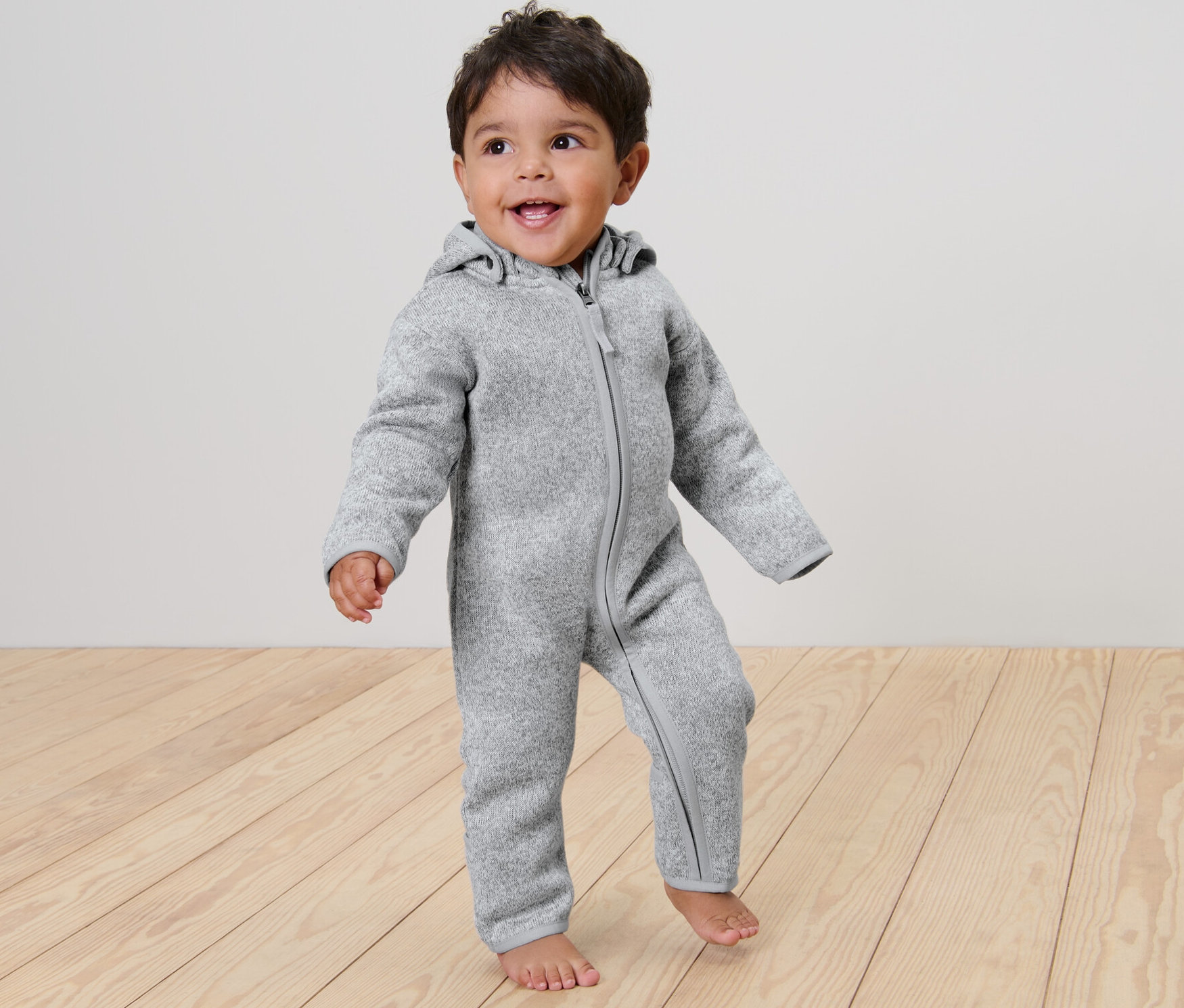 Un enfant souriant se tient sur un plancher en bois et porte une combinaison en polaire tricotée grise pour bébé.