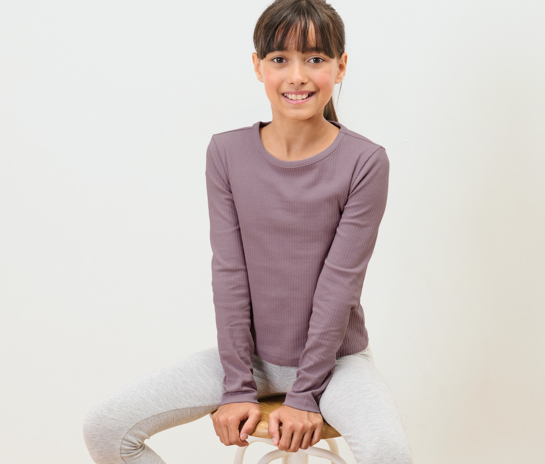 Une fille est assise sur un tabouret et porte un t-shirt à manches longues pour enfant avec structure côtelée.