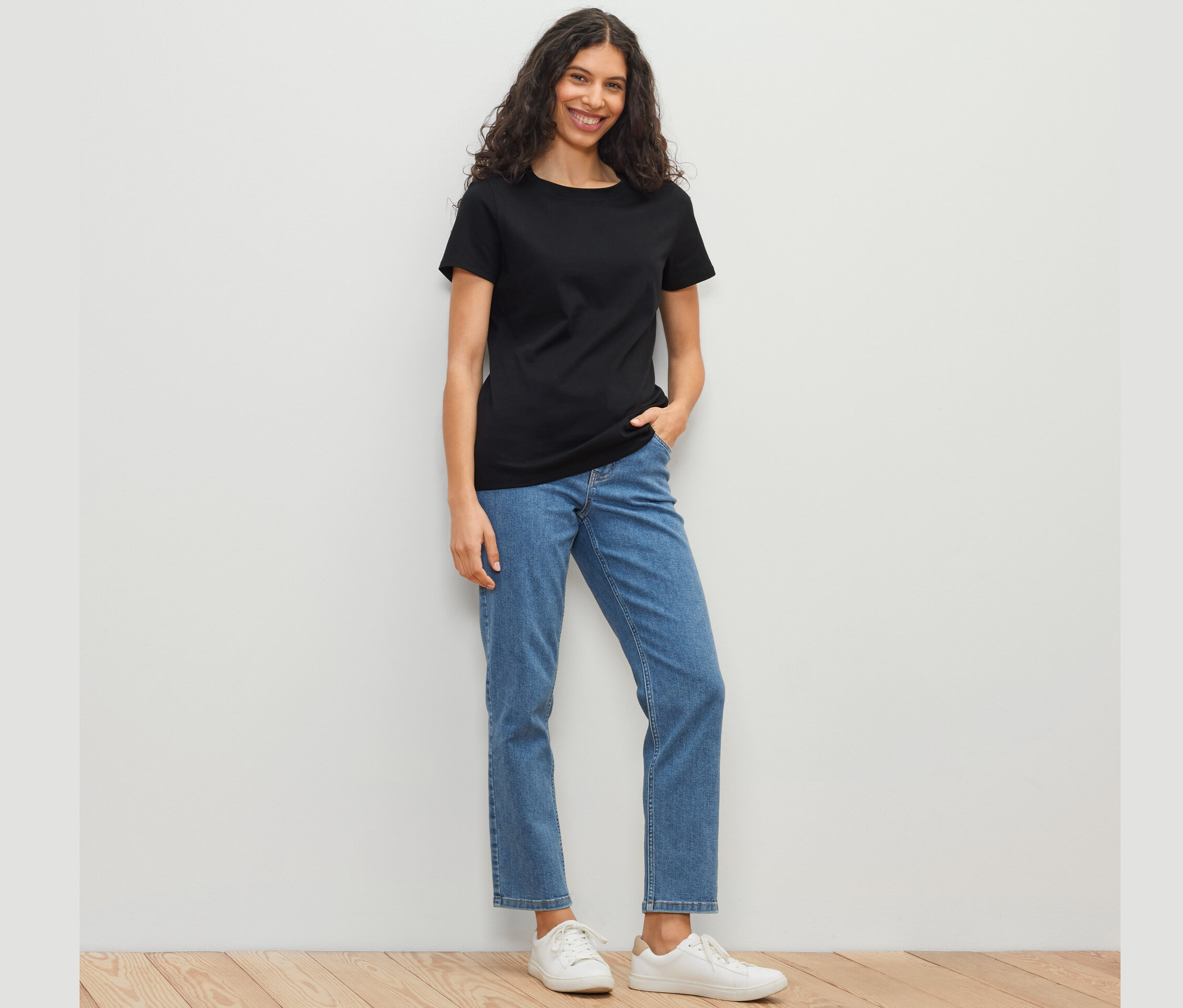 Femme portant un t-shirt basique noir, un jean bleu et des chaussures blanches debout devant un mur blanc.