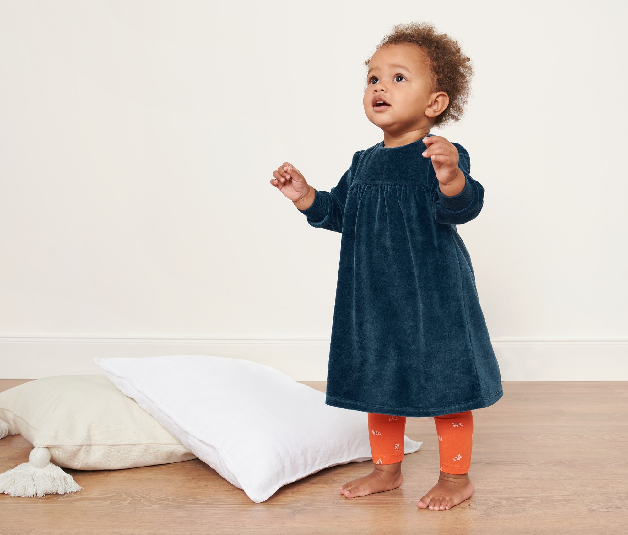 Un bébé se tient sur le sol à côté des coussins et porte une robe nicki pour bébé.