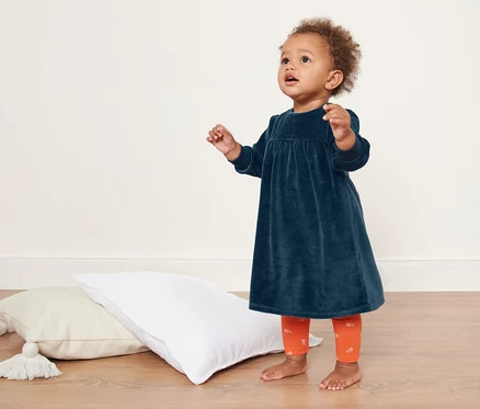 Un bébé se tient sur le sol à côté des coussins et porte une robe nicki pour bébé.
