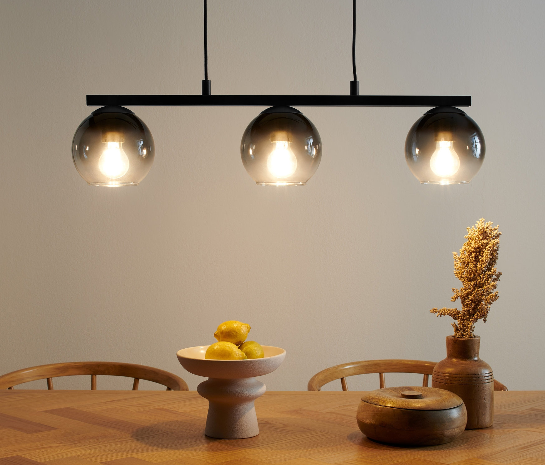 Lampes à suspension avec abat-jours en verre fumé au-dessus d'une table en bois. Sur la table, il y a une coupe de fruits et une boîte en bois.