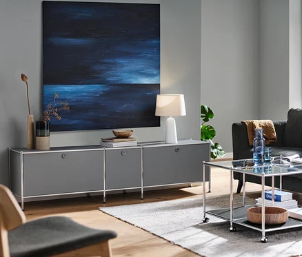 Meuble bas gris « CN3 » avec 3 abattants, une lampe de table avec abat-jour blanc et un canapé 3 places anthracite dans un salon. Egalement une table basse à roulettes « CN3 » et une chaise d'intérieur.